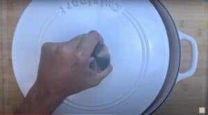 Hand lifting lid of white Dutch oven on wooden surface. Cooking preparation in progress.